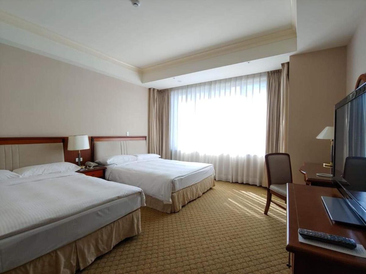 Evergreen Laurel Hotel - Тайчжун Экстерьер фото The image shows a hotel room featuring two single beds with white bedding. There is a large window with sheer curtains letting in natural light, and the walls are painted in neutral tones. A wooden desk with a chair is positioned near the window, and