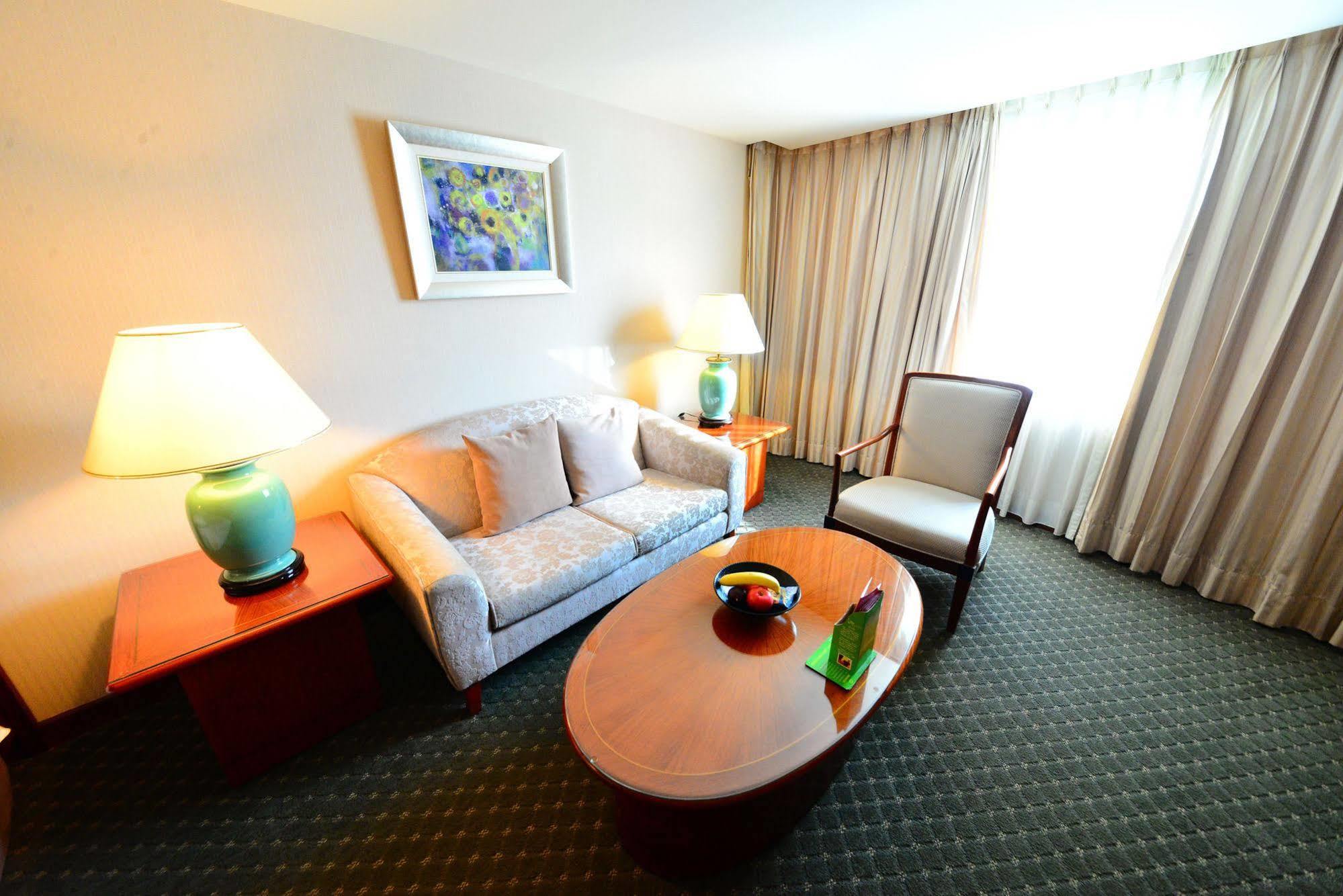 Evergreen Laurel Hotel - Тайчжун Экстерьер фото The photo shows a cozy living space, likely a hotel room or a lounge area. It features a light-colored sofa with cushions, a wooden coffee table in the center, and a comfortable armchair beside it. There are two lamps with turquoise bases on side tab