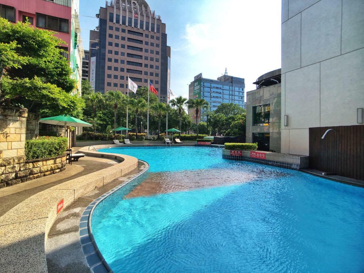 Evergreen Laurel Hotel - Тайчжун Экстерьер фото The photo shows a swimming pool set in a modern urban environment. The pool has a curved edge and is surrounded by greenery, including trees and umbrellas for shade. In the background, there are several tall buildings, indicating a cityscape, with so