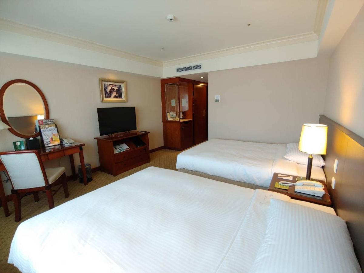 Evergreen Laurel Hotel - Тайчжун Экстерьер фото The photo shows a hotel room with two beds, neatly made with white linens. There is a desk and chair in one corner, accompanied by a flat-screen TV mounted on the wall. A large mirror and a wooden cabinet are also visible. The room is illuminated by 