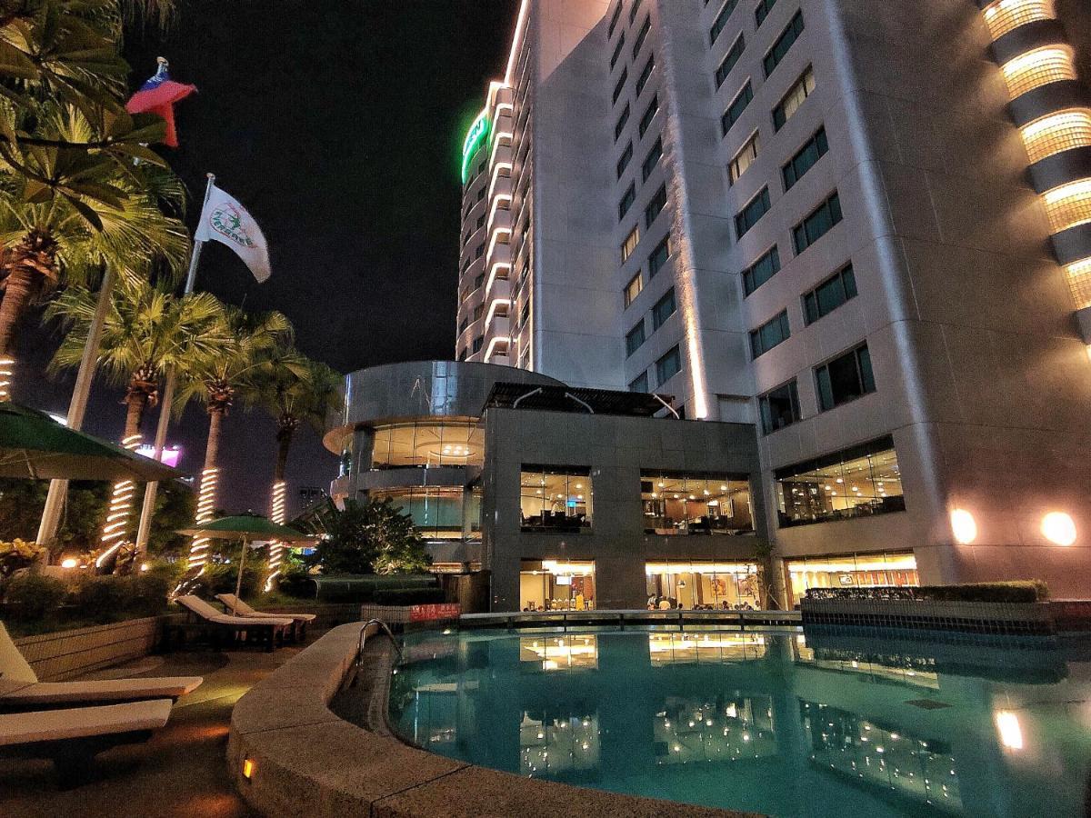Evergreen Laurel Hotel - Тайчжун Экстерьер фото The photo depicts a modern hotel or building at night, featuring a sleek facade illuminated by lights. In the foreground, there is a swimming pool that reflects the surrounding lights and palm trees. There are lounge chairs near the pool, and the atm